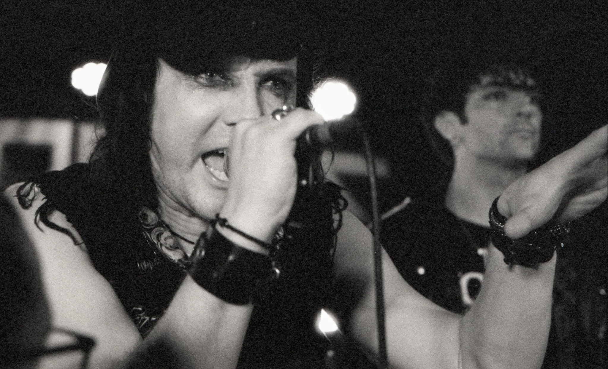 Black and white photo of a woman singing into a microphone with a man smiling in the background.